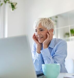 A woman with grey hair and a blue shirt sitting at a table looking at a laptop computer.